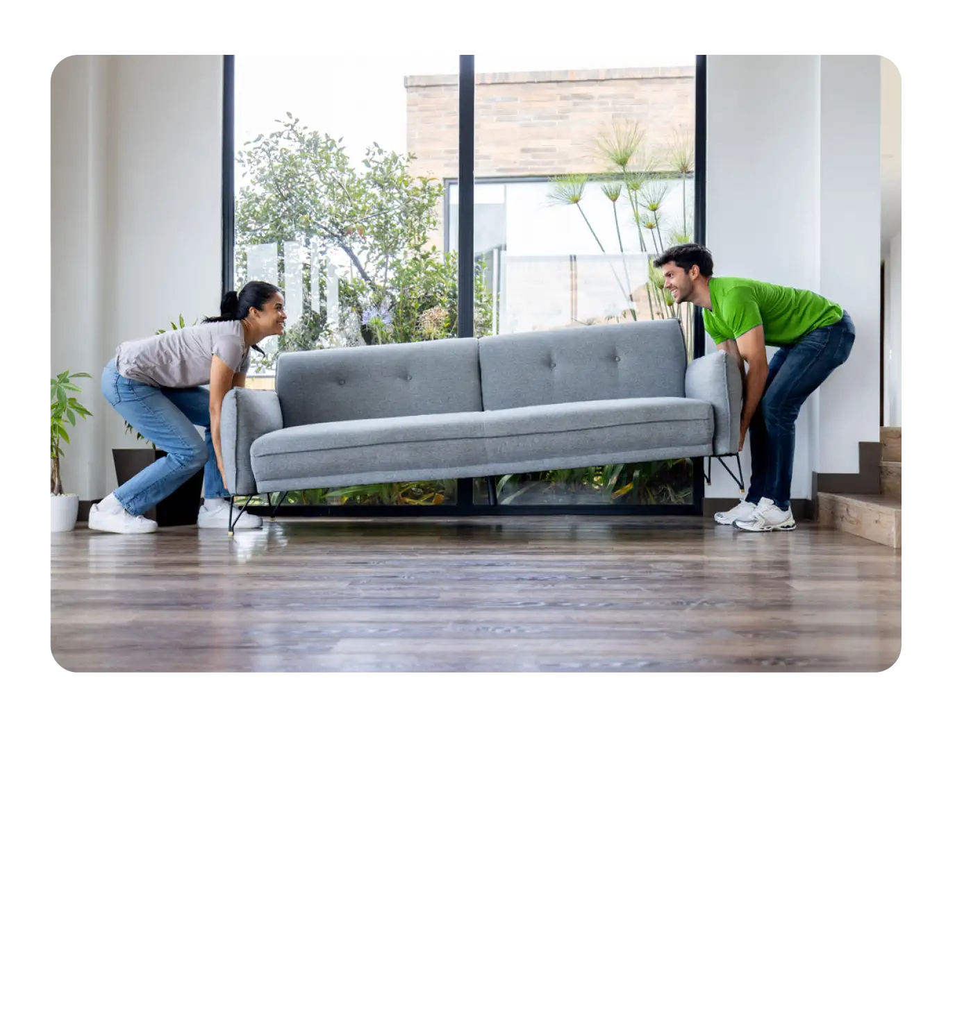 A couple moving a grey couch in a bright, modern living space with large windows.