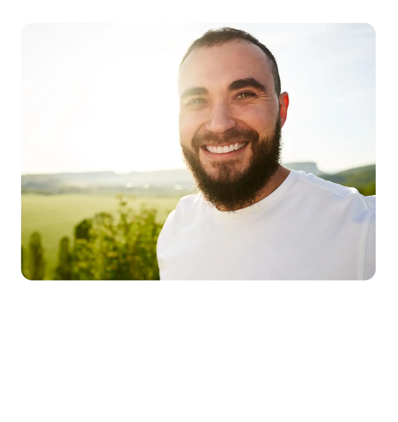 A smiling bearded man in a white t-shirt outdoors with a bright green scenic landscape in the background.
